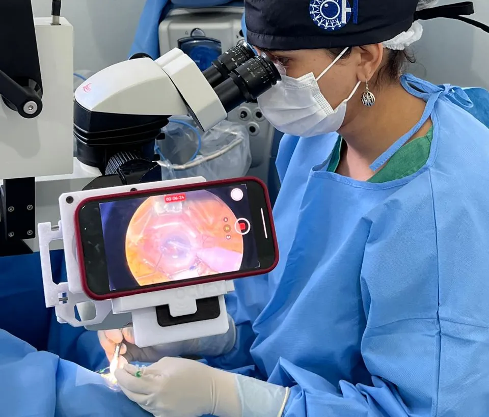 Cirurgiã da equipe opera ao microscópio; o celular acoplado mostra a imagem do olho