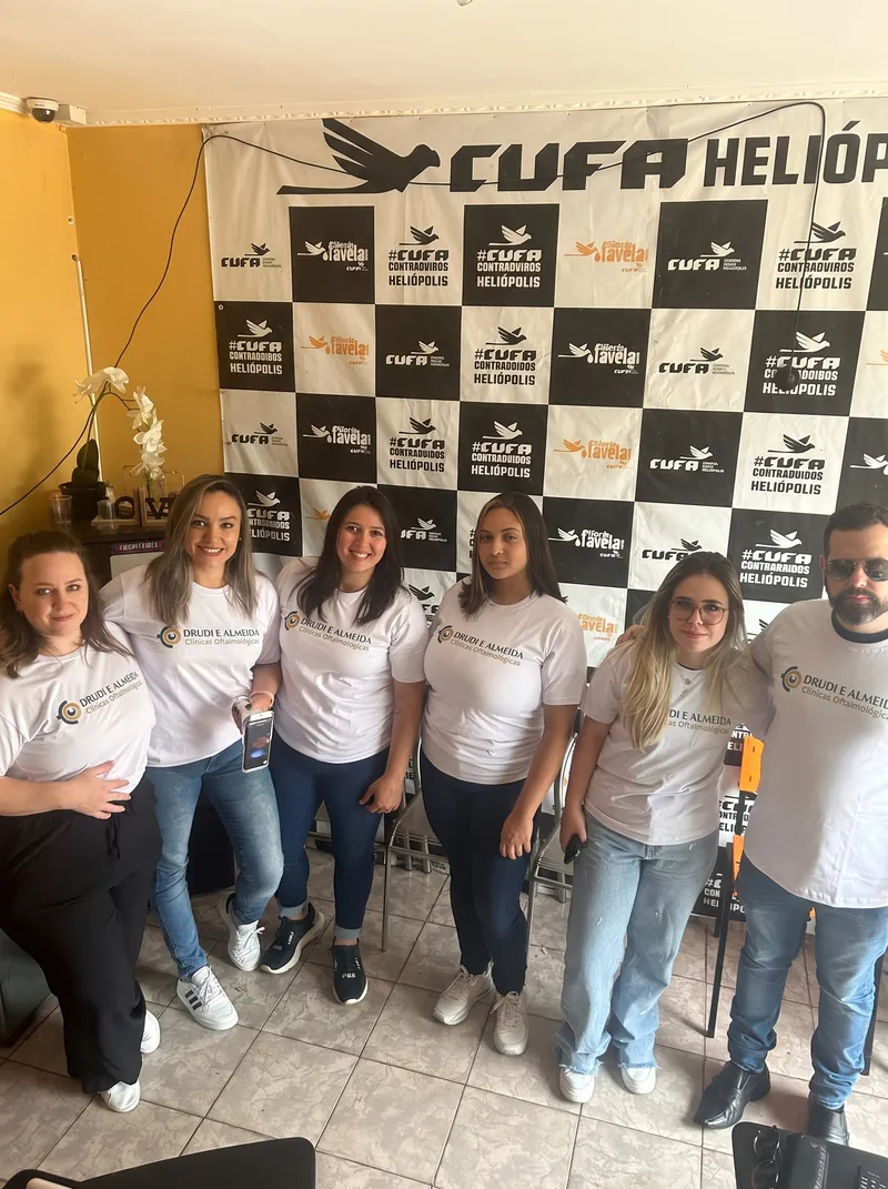 Seis pessoas da equipe, de camiseta branca com o logotipo da rede, diante do painel da CUFA Heliópolis