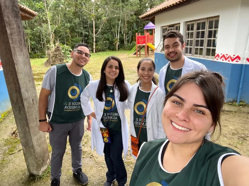Cinco pessoas da equipe, de jaleco e colete verde com o logotipo da rede, no pátio da escola da aldeia